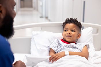 African american male doctor talking to boy patient in hospital bed in corridor with copy space. Hospital, medical and healthcare services.